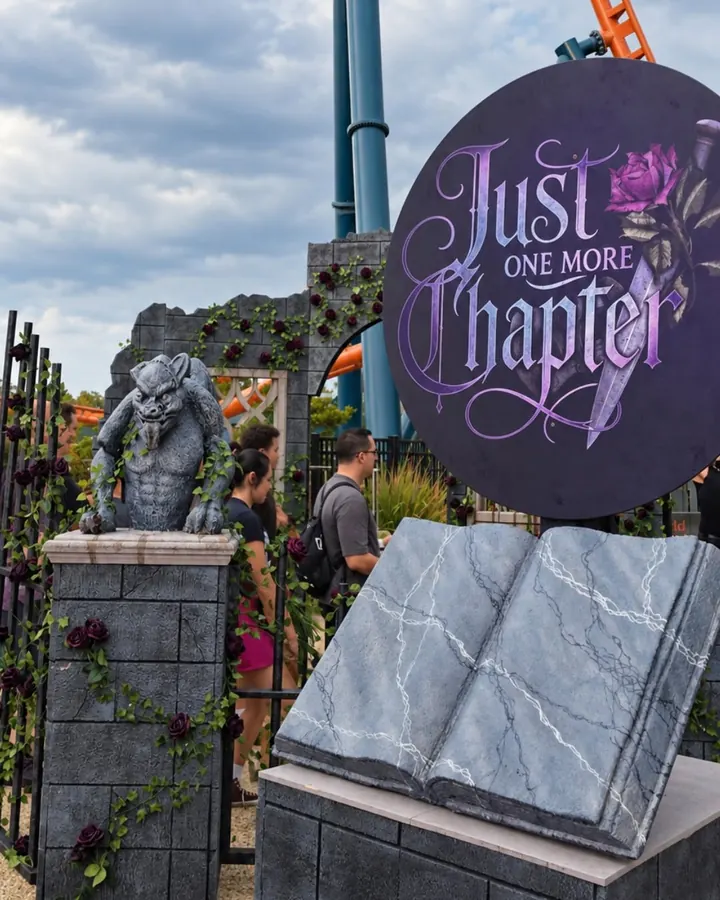 The purple Just One More Chapter sign above a stone book, next to a gargoyle on a rose-covered pillar