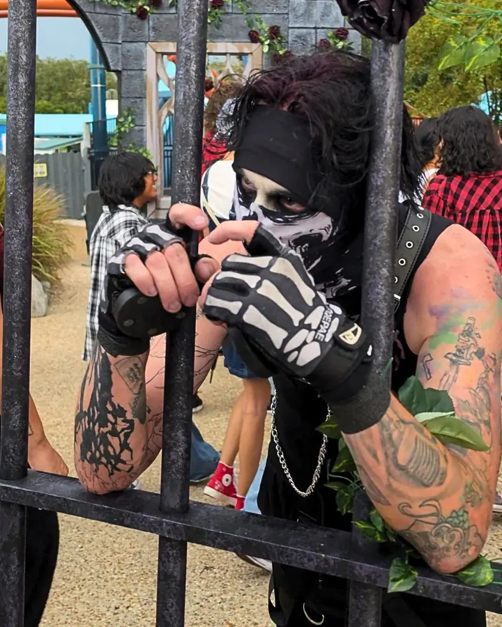 A tattooed performer in a skull mask and skeleton gloves leaning through the bars of an iron gate