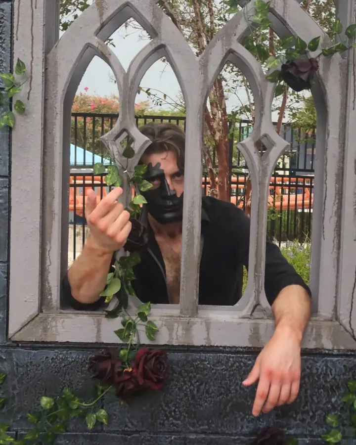 A performer in a black half mask beckoning through a gothic stone window wrapped in ivy and dark red roses